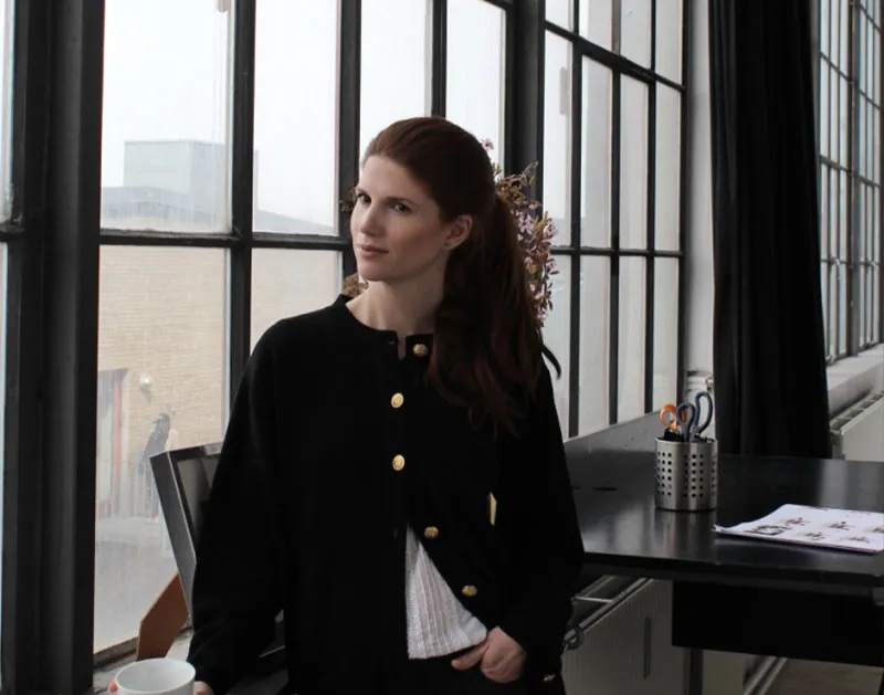 A woman leaning on a desk by the tall windows of an office